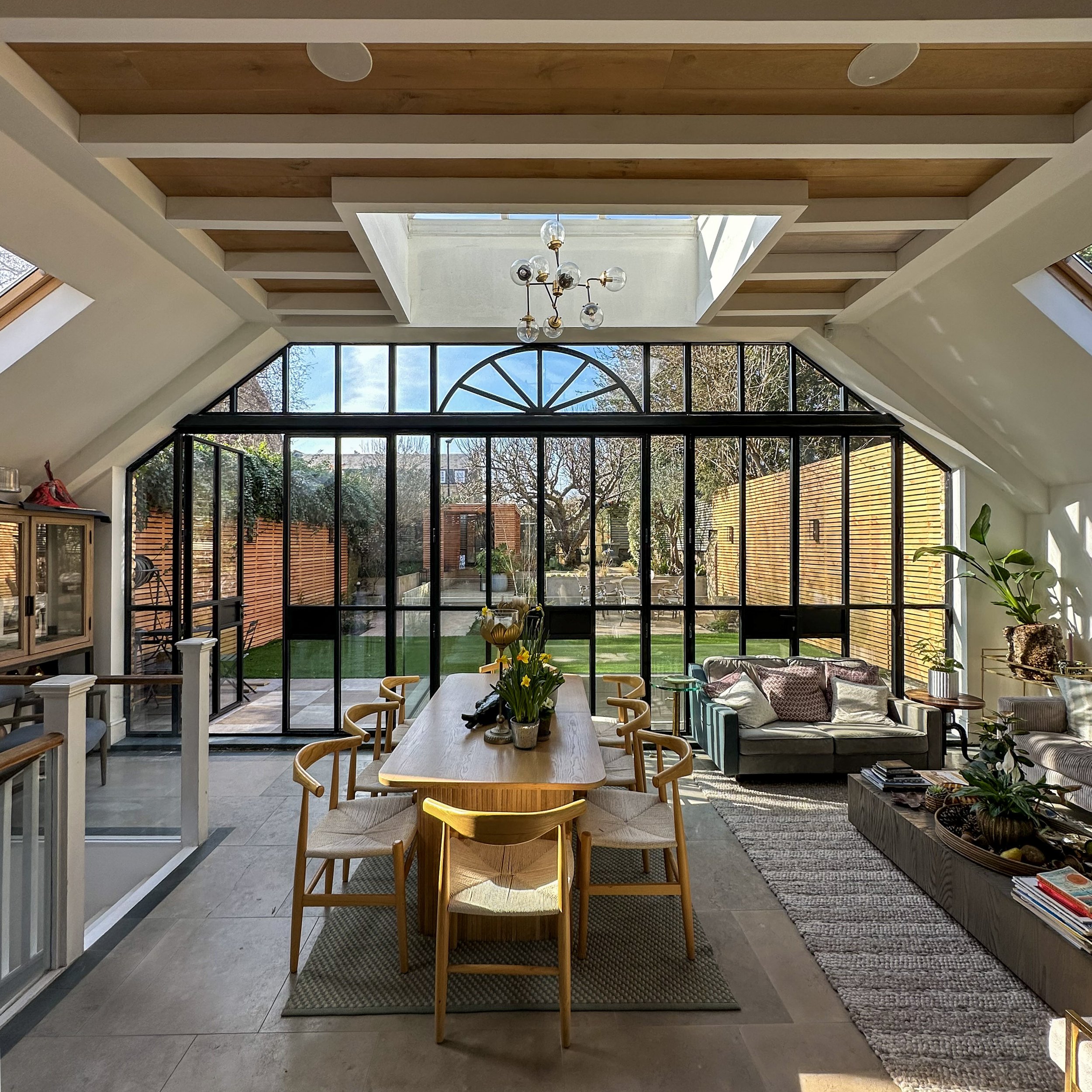 A modern dining room with a long wooden table surrounded by chairs, featuring a skylight and a chandelier above. The room has large glass doors and windows leading to a garden, with abundant natural light. A cozy seating area with sofas and plants is visible on the right.